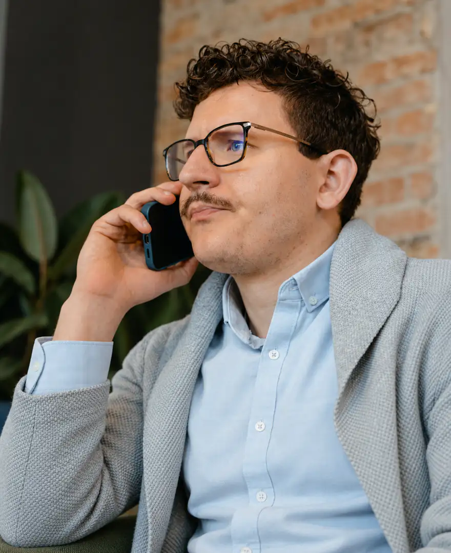A man in glasses on a phone call, seated by a brick wall