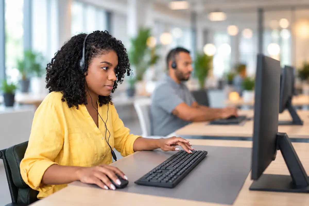 A call center agent in a headset working at her computer, a colleague on a call behind her